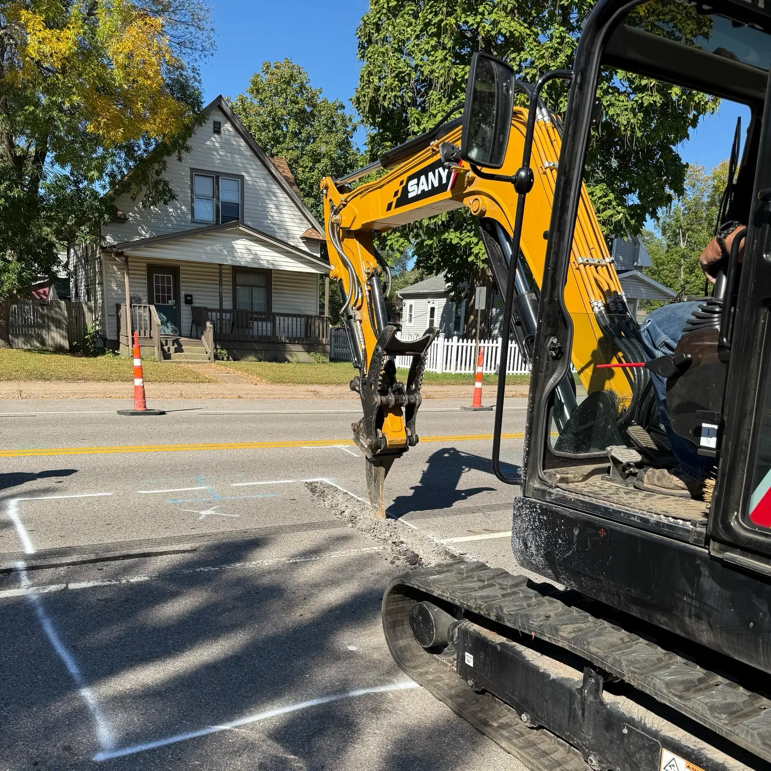 Excavator digging on street for sewer line replacement, with nearby residential houses and safety cones visible.