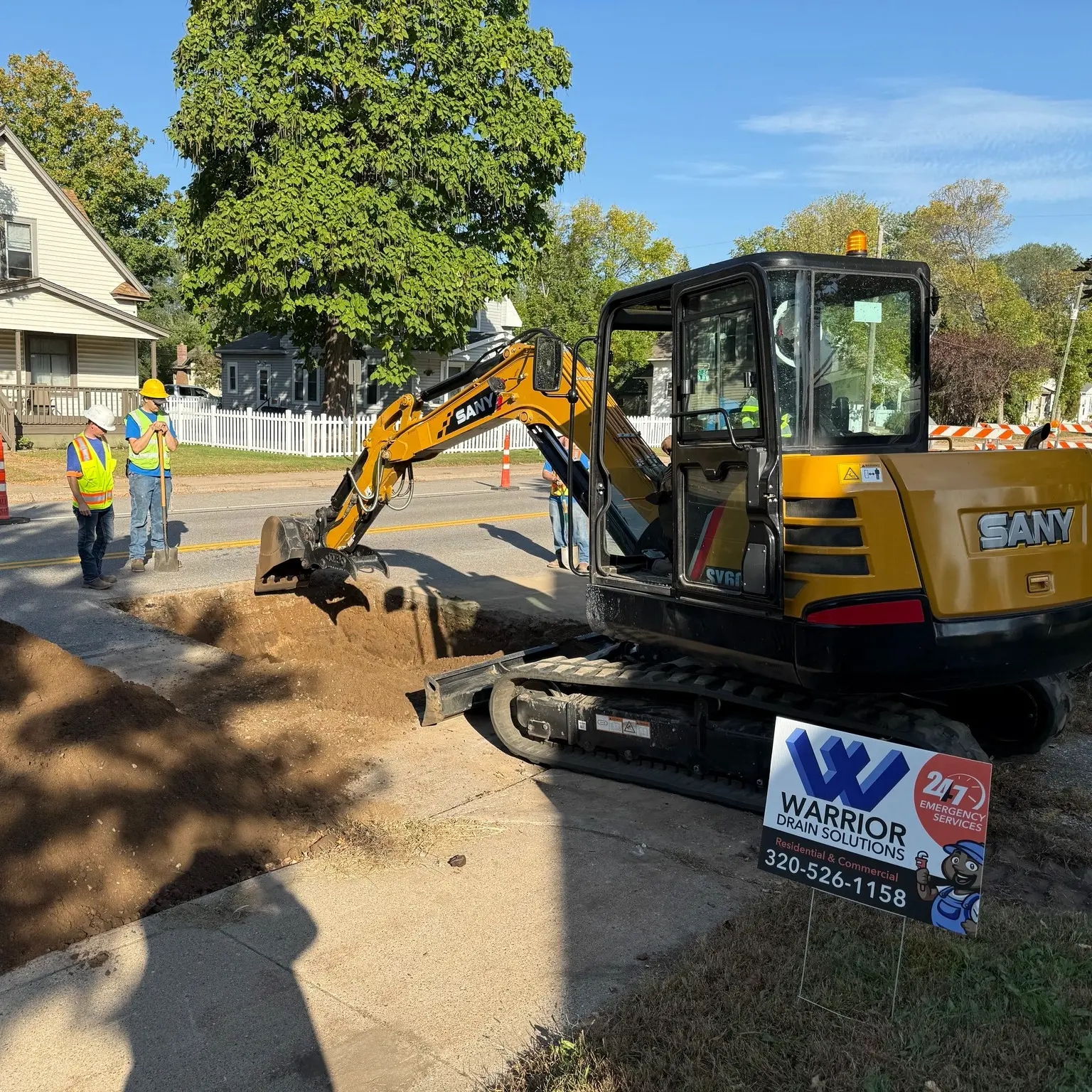 Excavator digging a trench for deep sewer line replacement, with workers in safety vests overseeing the operation, and a sign for Warrior Drain Solutions prominently displayed.