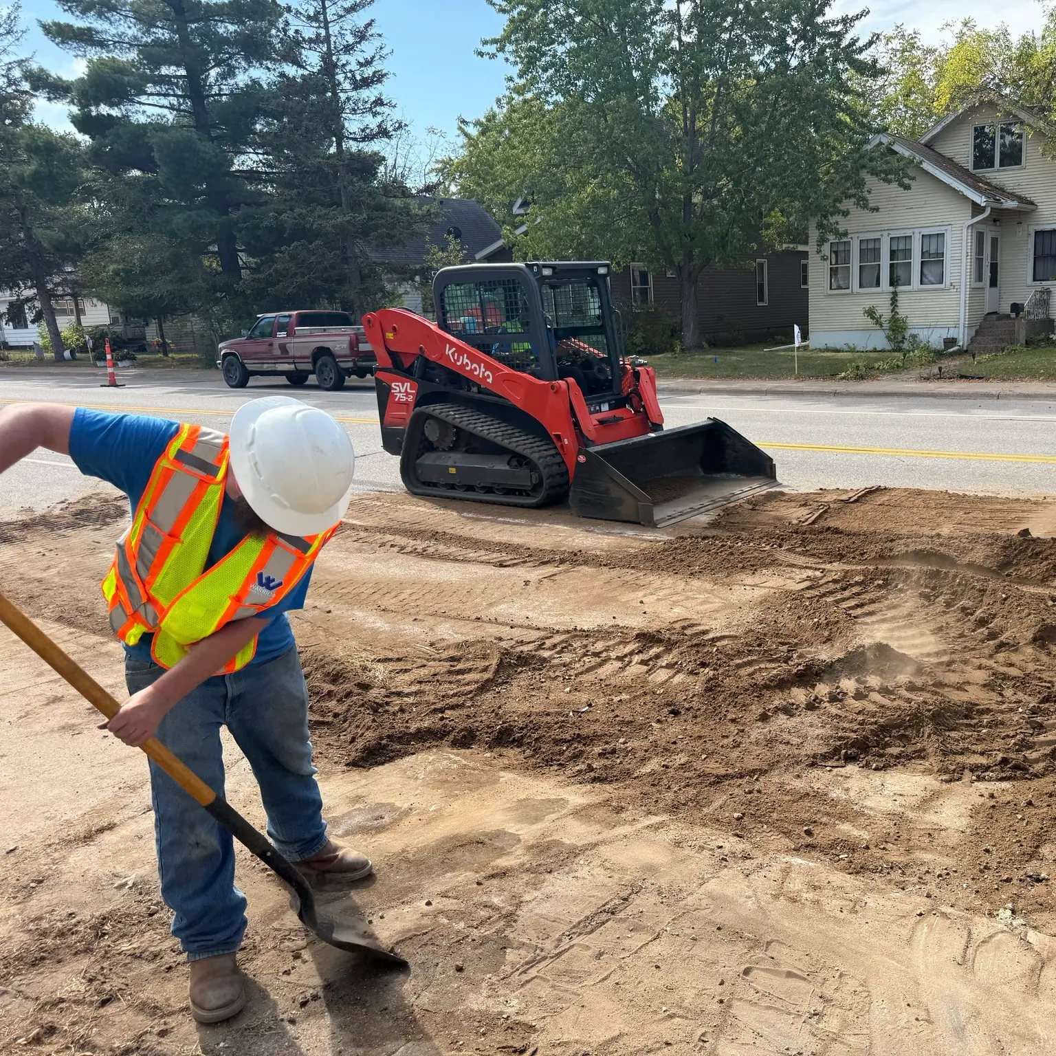 Construction worker in safety gear using a shovel on a trench site with a Kubota skid steer loader in the background, illustrating sewer line replacement project by Warrior Drain Cleaning & Plumbing Services.