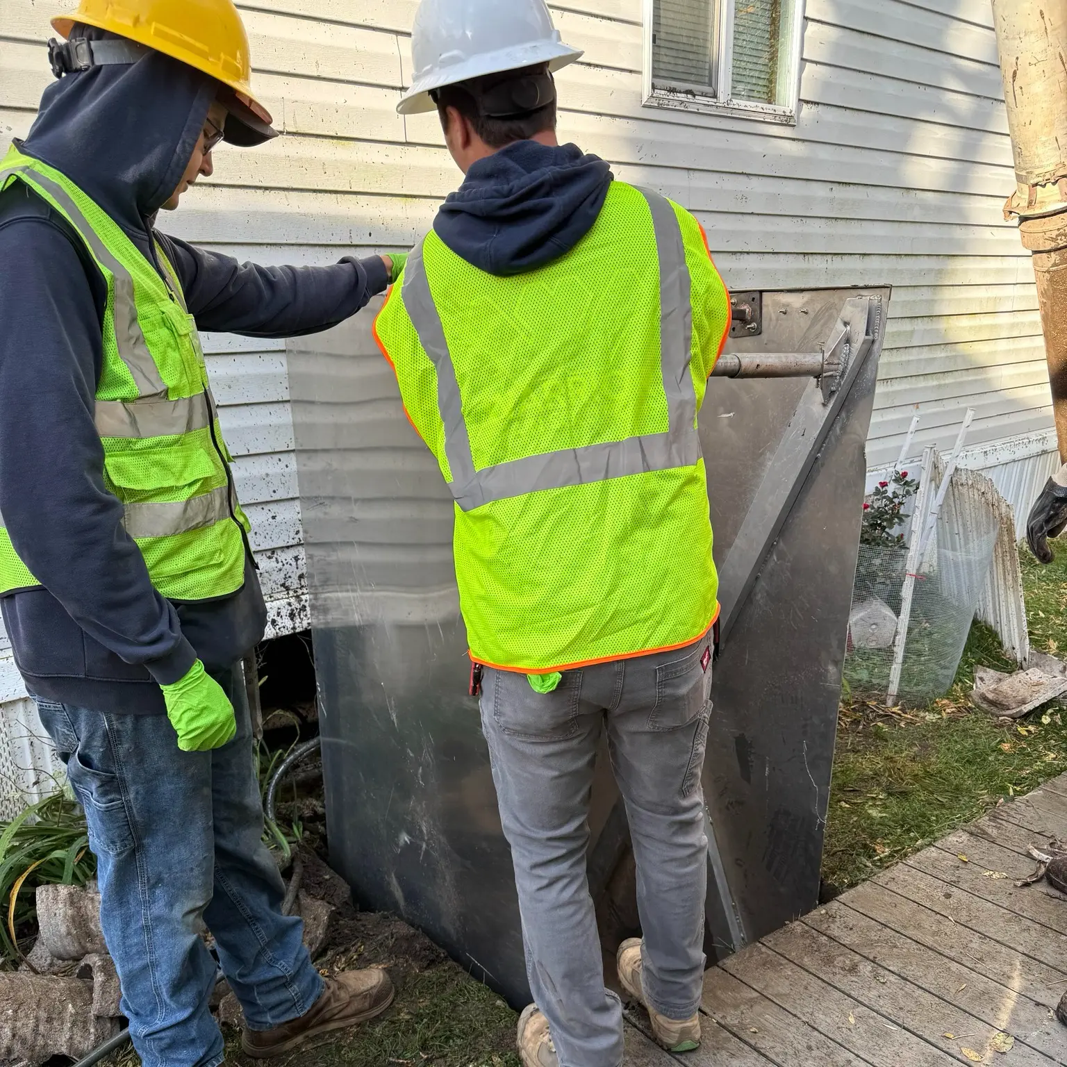 Two workers in safety vests inspecting a water system access panel, showcasing plumbing expertise in a residential setting.
