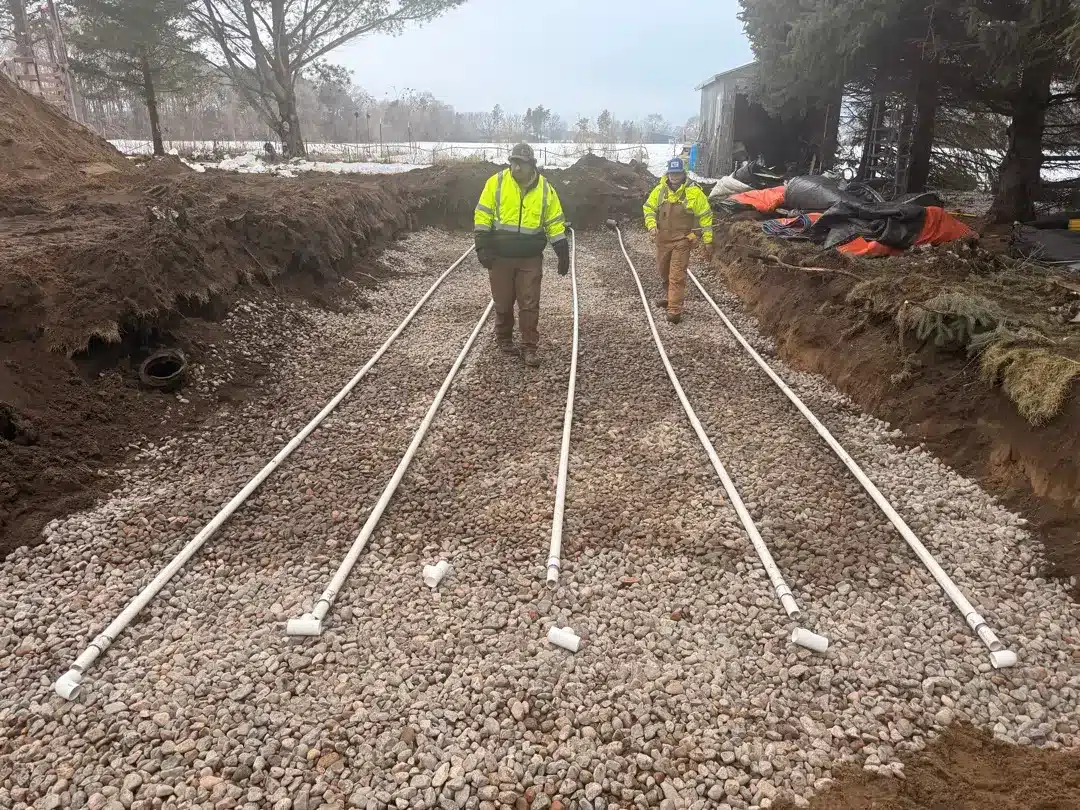 Workers installing a new septic system and drain field with PVC pipes on a gravel bed in cold weather conditions.