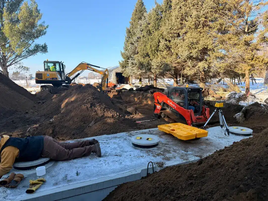 Septic system installation in winter, construction equipment, workers on site, and snowy landscape.