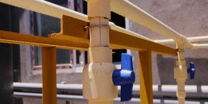 A off-white water pipe with a blue shut-off valve lever mounted outdoors on a yellow metal framework support.
