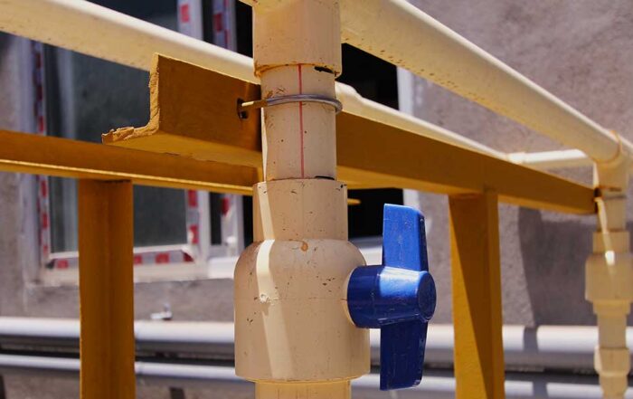 A off-white water pipe with a blue shut-off valve lever mounted outdoors on a yellow metal framework support.