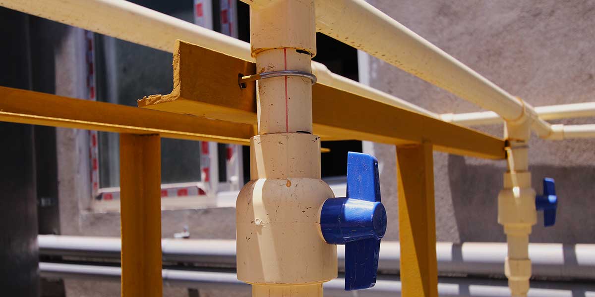A off-white water pipe with a blue shut-off valve lever mounted outdoors on a yellow metal framework support.